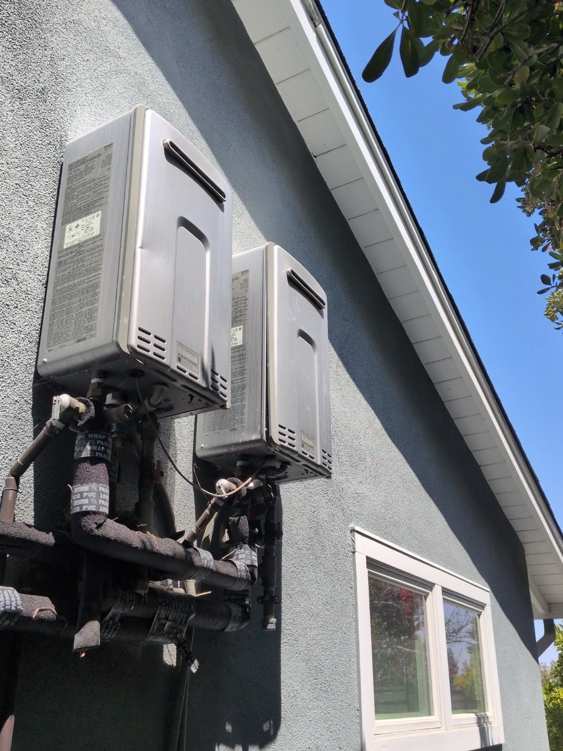 Before: two aging outdoor tankless water heaters with worn insulation and dated piping on a stucco exterior wall